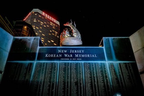 Night view of the New Jersey Korean War Memorial, featuring a wall of names and a sculpture, with Bally's Casino in the background.