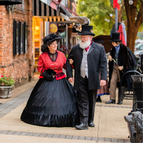 A woman in a red Victorian dress and a man in a black suit and top hat walk together on a historic street.
