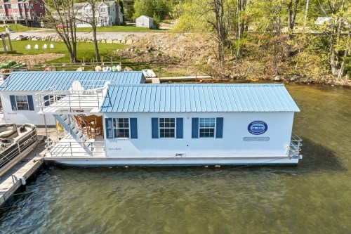 A houseboat with a blue metal roof docked on calm water, surrounded by trees and a grassy shoreline.