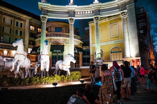 People gather outside Caesars Palace, admiring illuminated horse statues at night.
