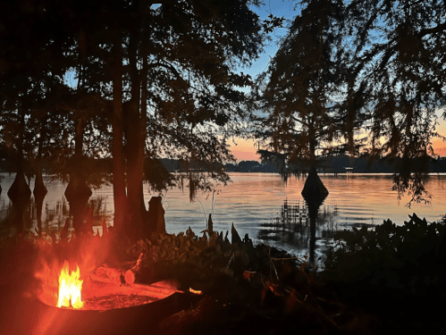 A serene lakeside scene at dusk, featuring a glowing campfire and silhouetted trees against a colorful sky.