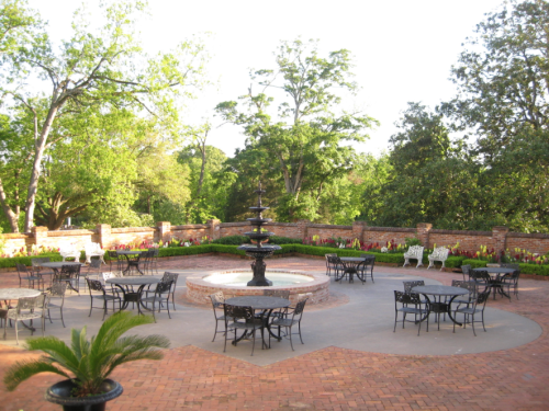 A serene outdoor courtyard with a fountain, surrounded by greenery and flower beds, featuring tables and chairs.