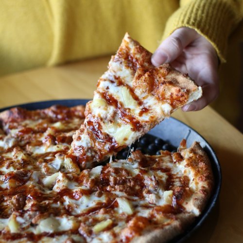 A hand lifts a slice of cheesy pizza from a round pan, with toppings visible and a cozy yellow sweater in the background.