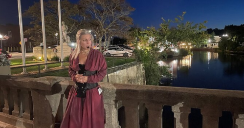 A person in a long maroon dress stands by a stone railing at night, with a calm river and lights in the background.