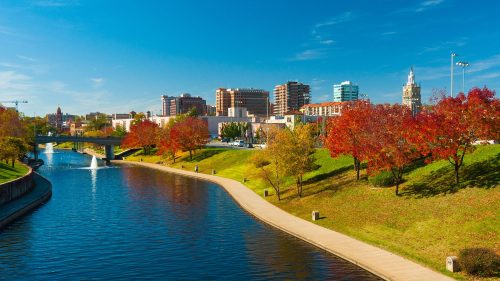 A scenic view of a city skyline with colorful autumn trees along a river under a clear blue sky.