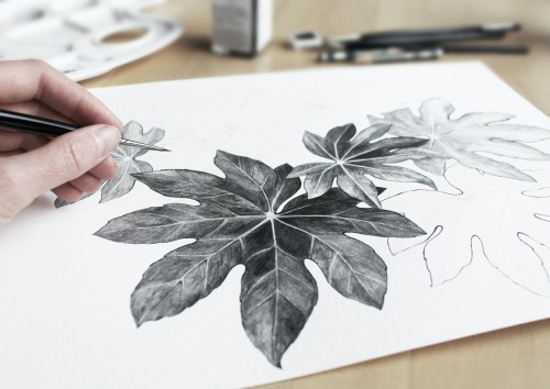 A hand drawing detailed leaves in pencil on a sketchpad, with art supplies in the background.