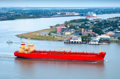 A large red cargo ship navigates a river, with buildings and another vessel visible in the background.