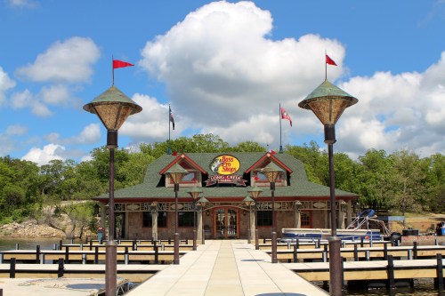 A waterfront building with a sign reading "Bass Shop" surrounded by docks and lampposts under a cloudy sky.