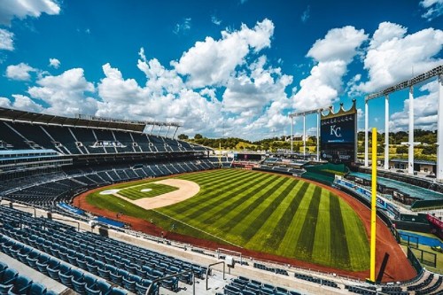 A wide view of a baseball stadium with a well-maintained field and blue sky filled with fluffy clouds.