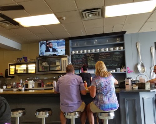 A casual diner scene with patrons sitting at the counter, watching a TV, and a chalkboard menu in the background.