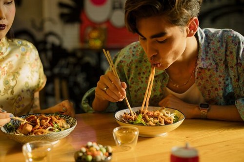 A young man eats noodles with chopsticks while a woman enjoys a dish at a table, surrounded by colorful decor.