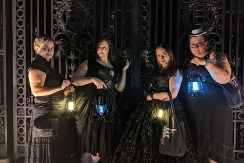 Four women in dark clothing hold lanterns in front of an ornate gate, creating a mysterious and gothic atmosphere.