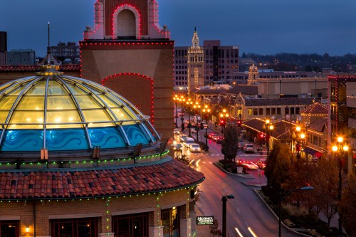 A cityscape at dusk featuring a glass dome, illuminated buildings, and festive lights along a busy street.
