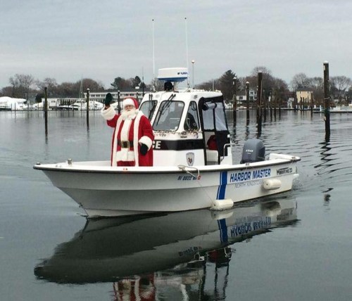 Santa Claus waves from a small boat labeled "Harbor Master" on a calm waterway, surrounded by winter scenery.