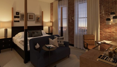 Cozy hotel room featuring a bed, sofa, desk, and large windows with city views, accented by warm lighting and brick walls.