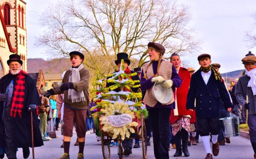 A festive parade with people in vintage clothing, carrying a decorated cart and celebrating in a lively atmosphere.