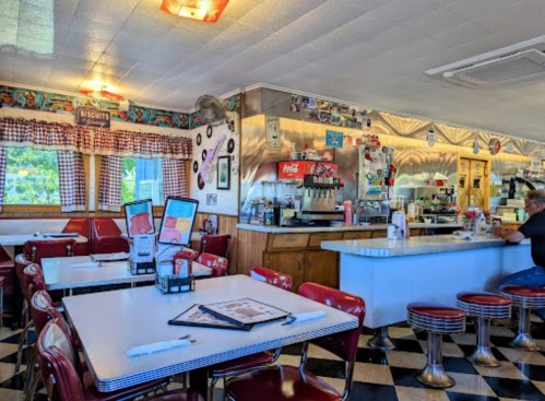 A retro diner interior with checkered floors, red booths, and a counter, featuring vintage decor and a customer at the bar.