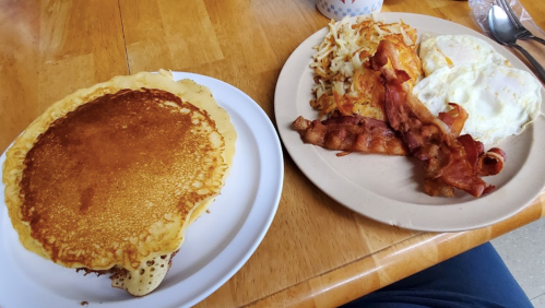 A plate with a large pancake beside another plate of eggs, bacon, and hash browns on a wooden table.