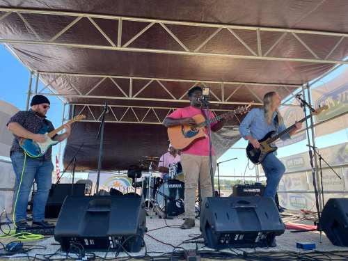 A band performs on stage with guitars and drums under a large canopy at an outdoor event.