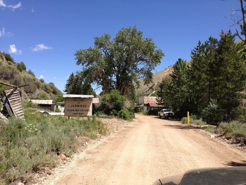 A dirt road leads to a small, rustic town sign for Jarbridge, surrounded by trees and mountains under a blue sky.
