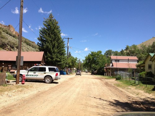 A dirt road lined with trees and buildings, featuring a sheriff's vehicle parked on the side under a clear blue sky.