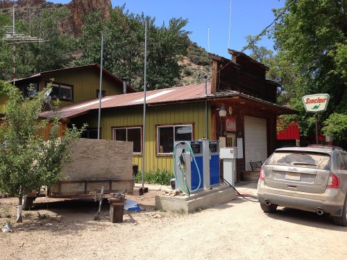 A rustic gas station with a green and yellow building, a fuel pump, and a parked car in a rural setting.