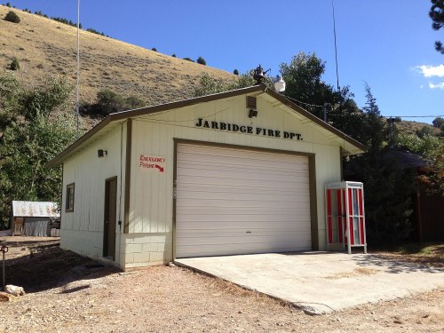 A green fire department building with a large garage door, surrounded by trees and mountains under a clear blue sky.