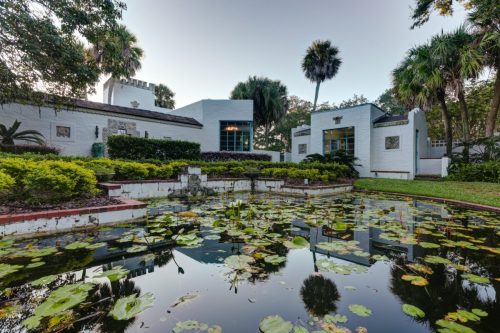 A serene garden with a pond covered in lily pads, surrounded by lush greenery and a modern white building in the background.