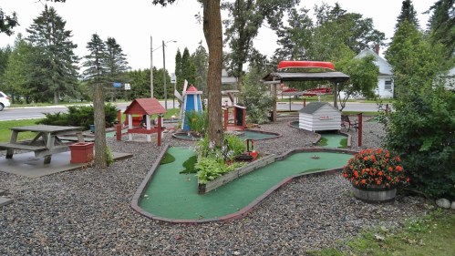 A miniature golf course with colorful structures, surrounded by trees and flowers, near a roadside.