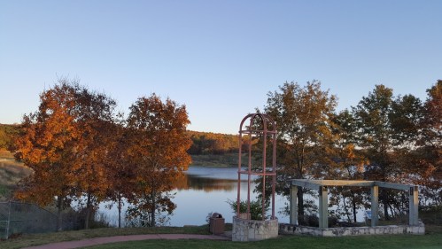 A serene lakeside view with autumn trees and a stone gazebo under a clear blue sky.