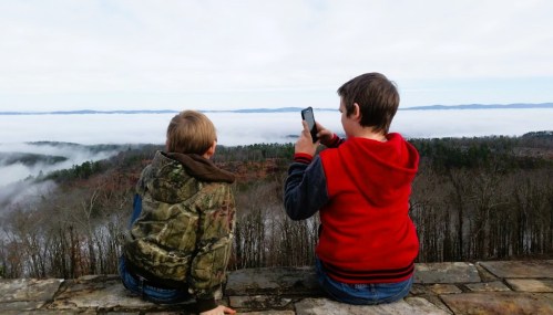 Two boys sit on a stone ledge, overlooking a foggy valley and distant mountains, one taking a photo with a phone.