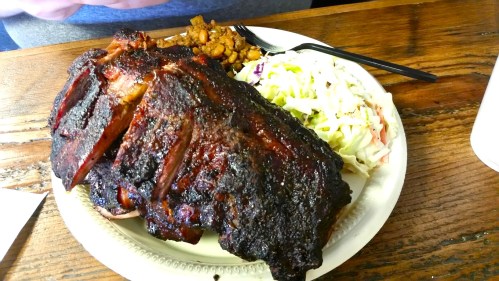 A plate featuring a large, smoked meat portion, coleslaw, and baked beans on a wooden table.