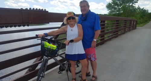 A smiling couple stands on a bridge with a bicycle, enjoying a sunny day by the water.