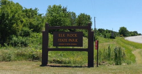 Sign for Elk Rock State Park, surrounded by greenery, with a clear blue sky and a road in the background.