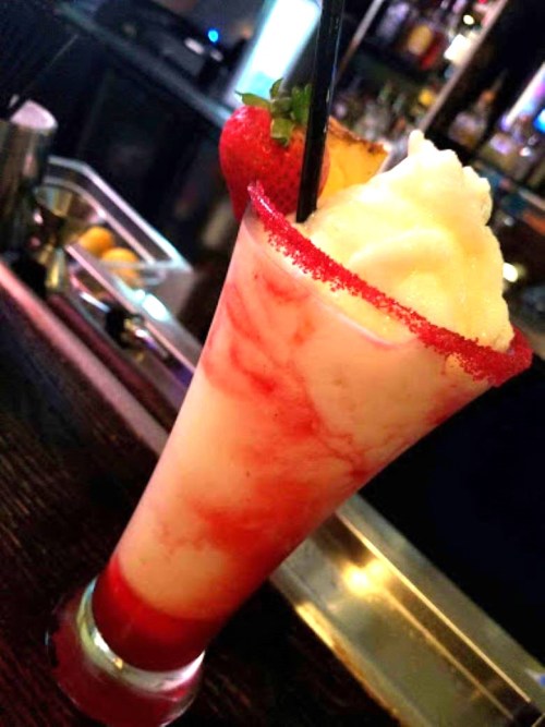 A colorful cocktail with a frosty top, garnished with a strawberry and pineapple, served in a tall glass.