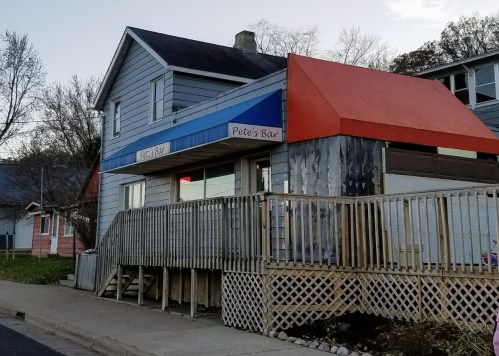 Exterior of Pete's Bar, a two-story building with a blue and orange awning, wooden deck, and nearby residential homes.