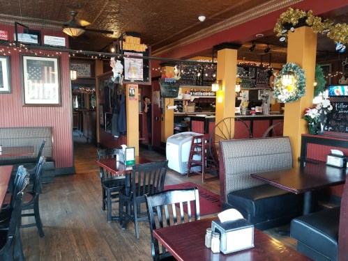 Cozy restaurant interior with wooden tables, chairs, and festive decorations, featuring a bar area in the background.