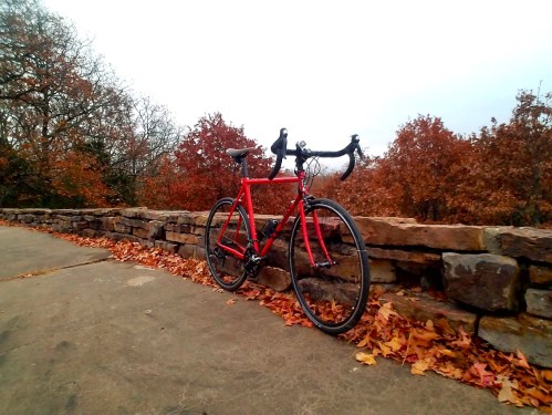 A red bicycle rests on a stone wall surrounded by autumn leaves and trees with orange foliage.