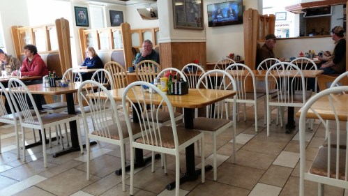 A cozy diner interior with wooden tables, white chairs, and patrons seated at various tables, some watching a TV.