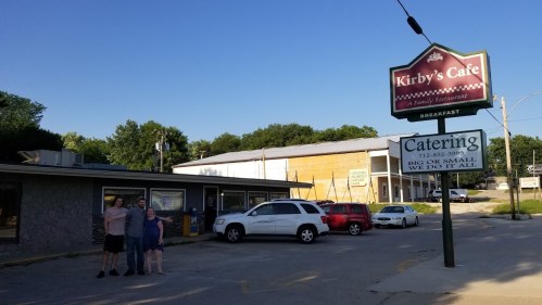 A family restaurant named Kirby's Cafe with a sign, featuring a few people standing outside and parked cars.