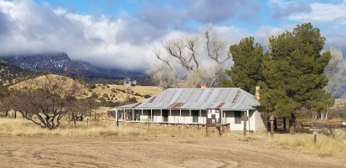 A rustic, weathered house with a metal roof, surrounded by trees and mountains under a cloudy sky.