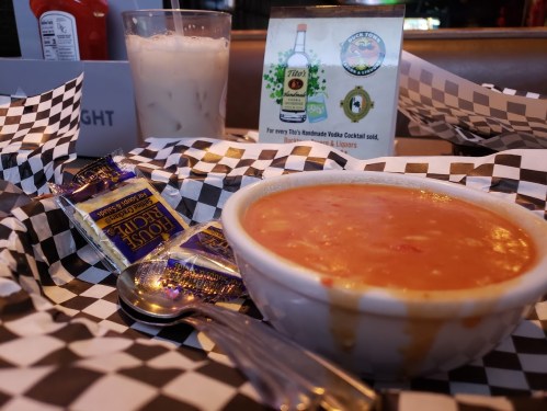 A bowl of soup with a spoon, condiments, and a drink in a restaurant setting, all on a checkered tablecloth.
