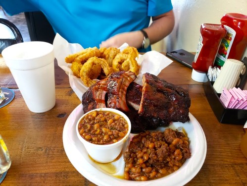 A plate of barbecue ribs, baked beans, and onion rings, with a drink and condiments in the background.