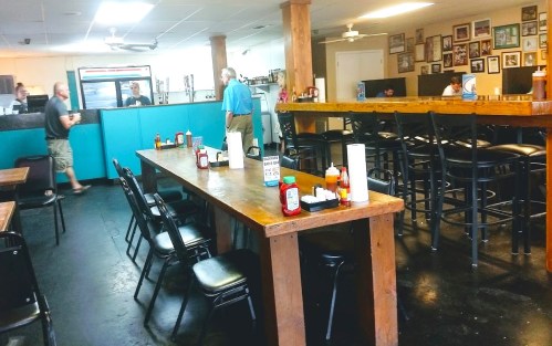A casual diner interior with wooden tables, black chairs, and a counter. Customers are seated and ordering at the counter.