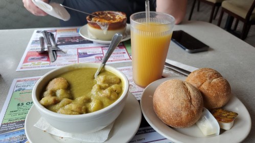 A bowl of green soup with dumplings, two rolls with butter, and a glass of orange juice on a table with a menu.
