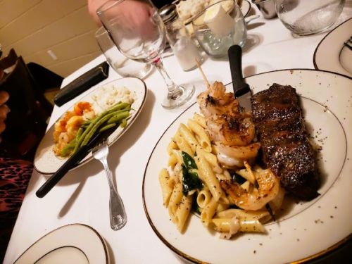 A plate with grilled shrimp, steak, pasta, and spinach, alongside a side of green beans and rice on a dining table.