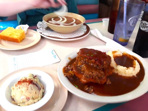A plate of meatloaf with gravy, mashed potatoes, and a side of coleslaw, with cornbread and a dessert in the background.