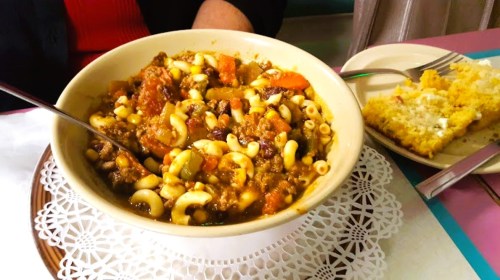 A bowl of hearty macaroni and beef stew with vegetables, alongside a plate of cornbread.