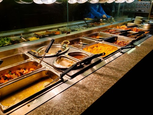 A buffet with various food trays, including meats, vegetables, and sauces, under warm lighting.