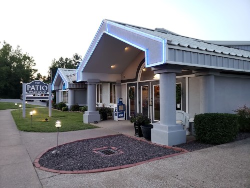 Exterior view of The Patio Steak House, featuring blue neon lights and a landscaped entrance at dusk.